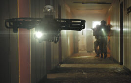 a drone flys down a dark hallway with outline of military troops carrying weapons in the background.